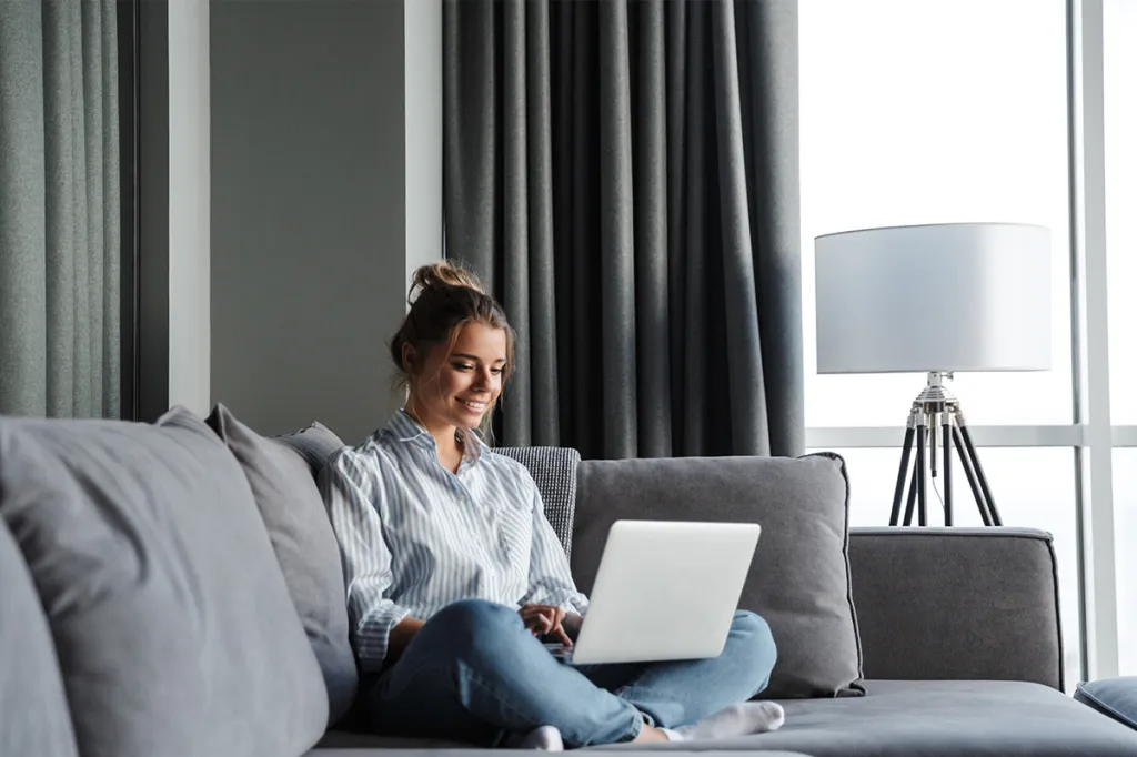 Woman sitting on a couch using a laptop in a bright apartment living room.